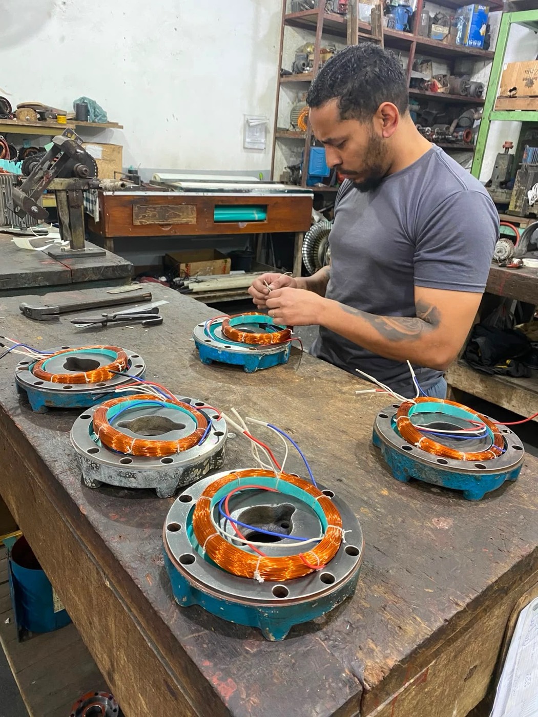 Técnico enrolando bobinas de cobre na bancada, com quatro estatores de motobomba em preparo simultâneo